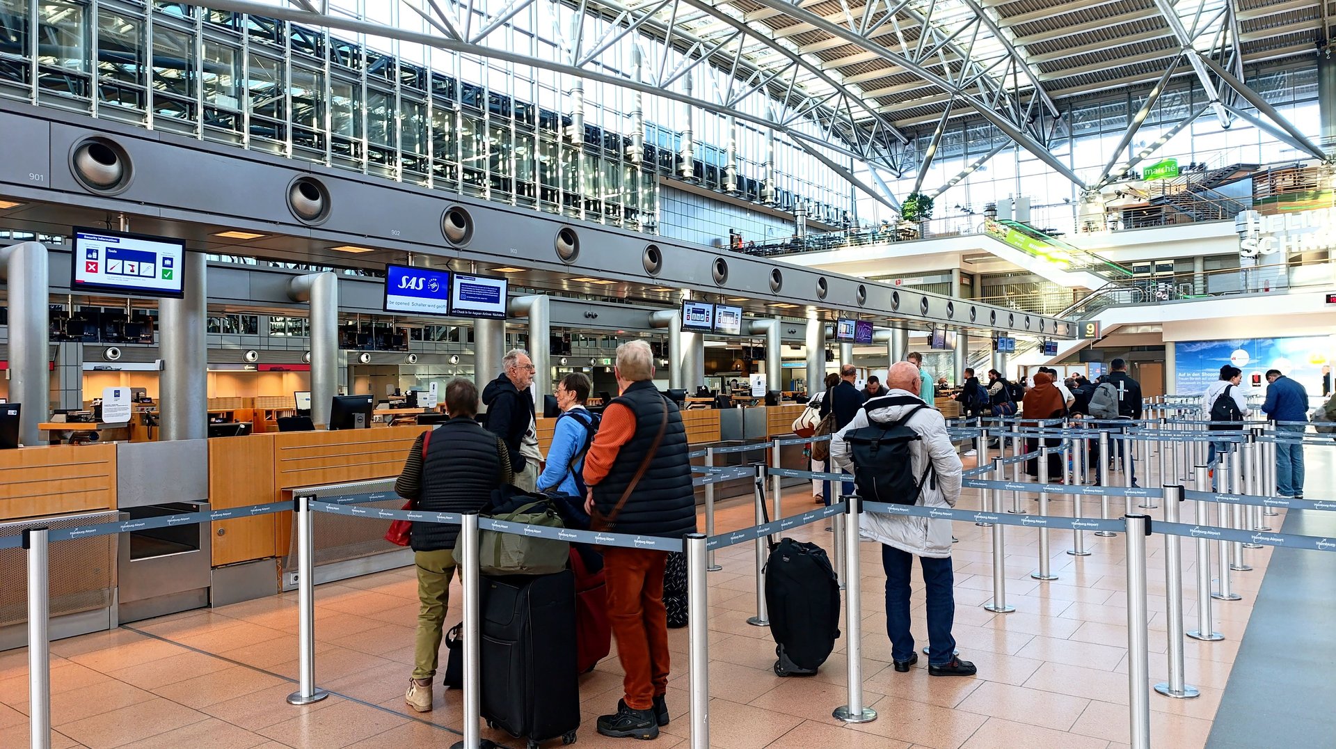 Pasajeros formados en filas con maletas en zona de check-in del aeropuerto de Hamburgo — counters europeos con múltiples carriles y pantallas de información sobre los mostradores